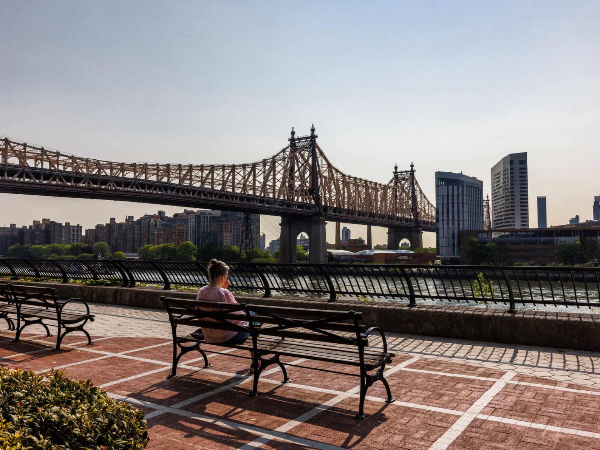 Woman sitting on a bench at Sutton Park looking out over the East River with the Queensboro Bridge in the distance.